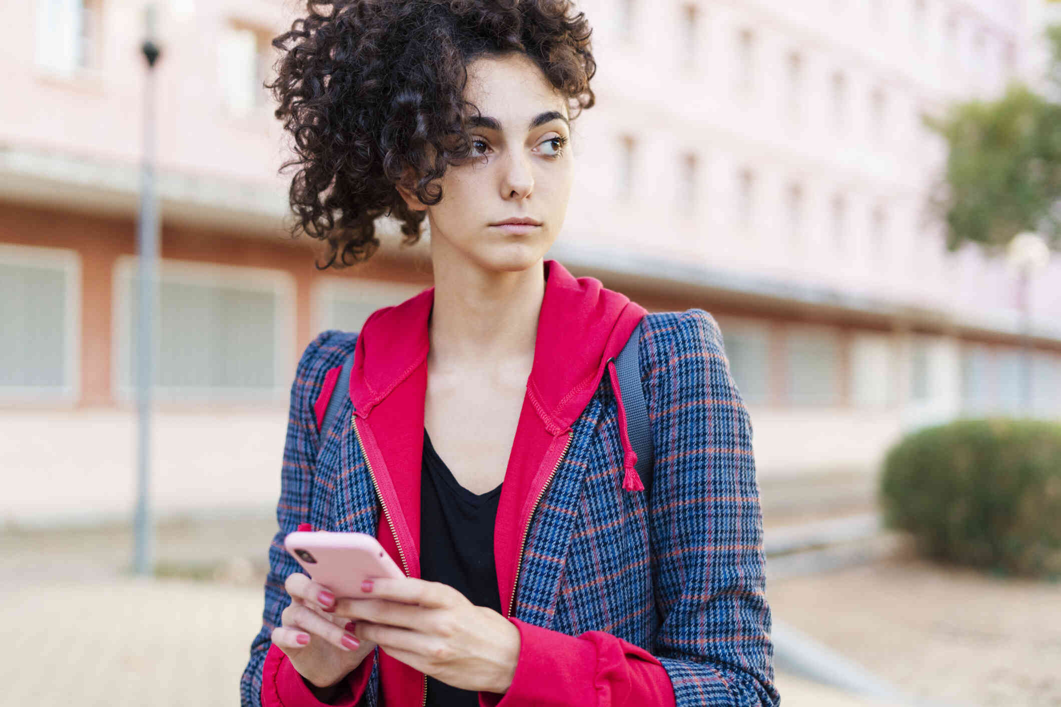 A woman ina blazer stands outside with her phone in her hands as she gazes off to the side with a serious expression.